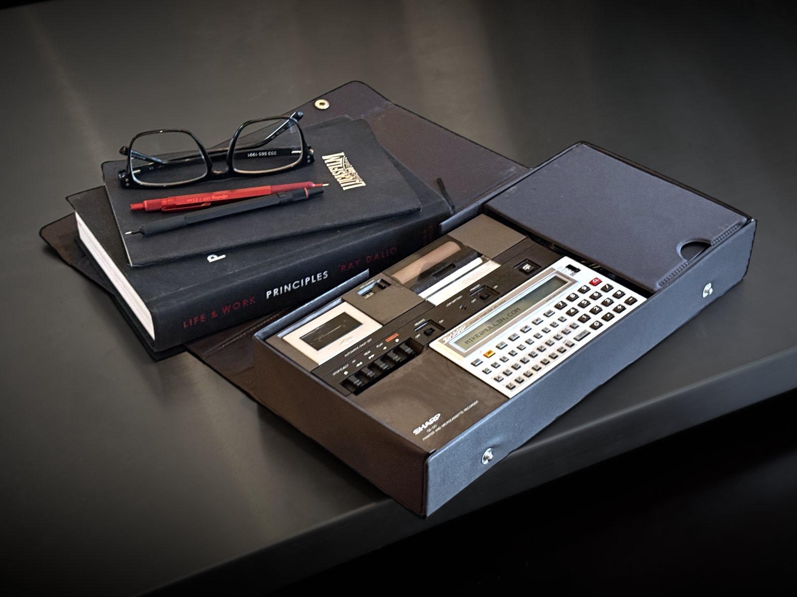 A Sharp PC-1250A pocket computer seated in its CE-125 printer and microcassette unit, in an open case on a steel counter beside glasses, pens and notebooks.